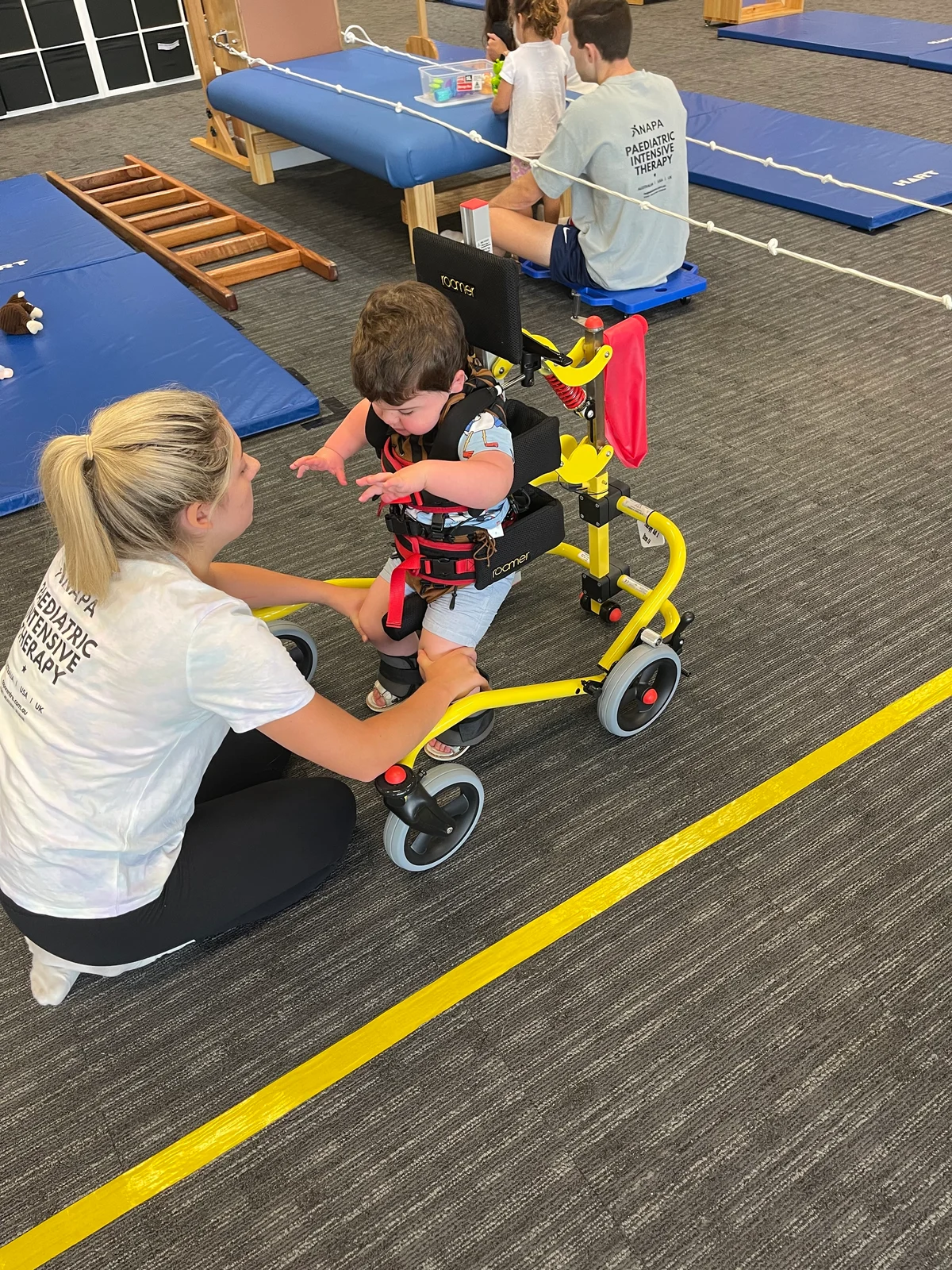 A young child using a supported walking frame during therapy with assistance from a clinician